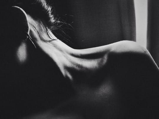 A black and white photo of a woman's naked, stretched fully to the side, showing her veins in her neck. Her hair is up and a few wisps catch the light. 