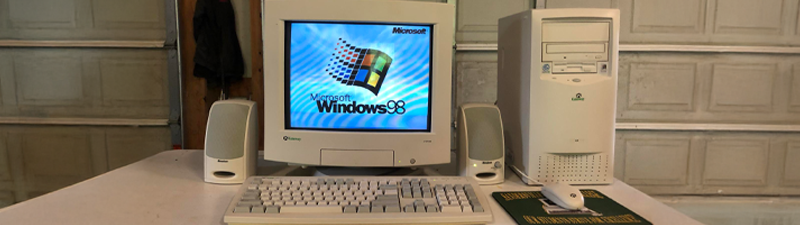 An old tower computer with a CRT screen sits on a desk in a dirty garage. It has an old keyboard, mouse, and speakers alongside it.