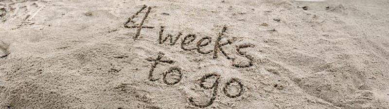 The words 'four weeks to go' are written in sand on a beach.