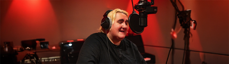 A blond-haired non-binary person sits in front of a microphone, wearing headphones. There are red lights in the background.