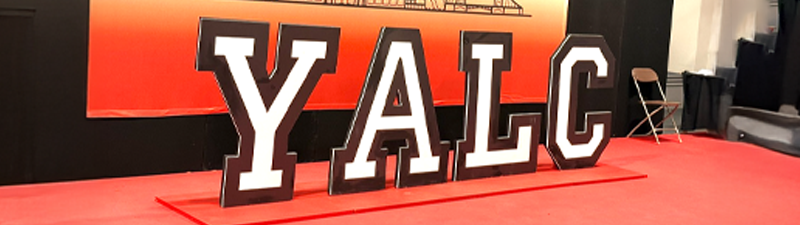 A photograph of the letters Y, A, L, and C cut out and standing up at last year's YALC event. They are white and black against a red backdrop.