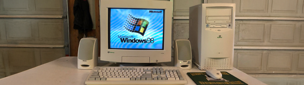 An old tower computer with a CRT screen sits on a desk in a dirty garage. It has an old keyboard, mouse, and speakers alongside it.