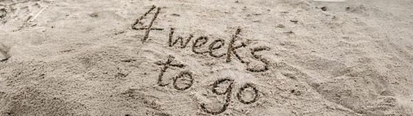 The words 'four weeks to go' are written in sand on a beach.