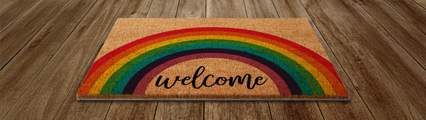 A welcome mat with a rainbow on it sits atop a wooden floor.