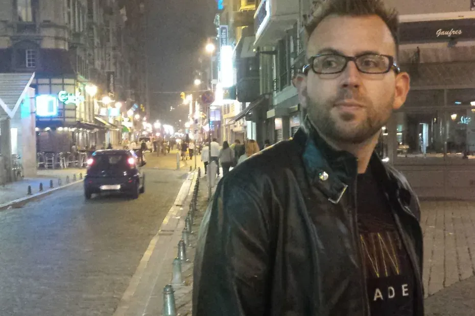 Me, a bespectacled person with stubble, wearing a black pleather jacket and Flynn's Arcade t-shirt, standing in a night-light street in Liège, Belgium