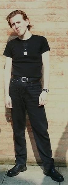 A teenager in all black, with dark hair, standing awkwardly in front of a brick wall.