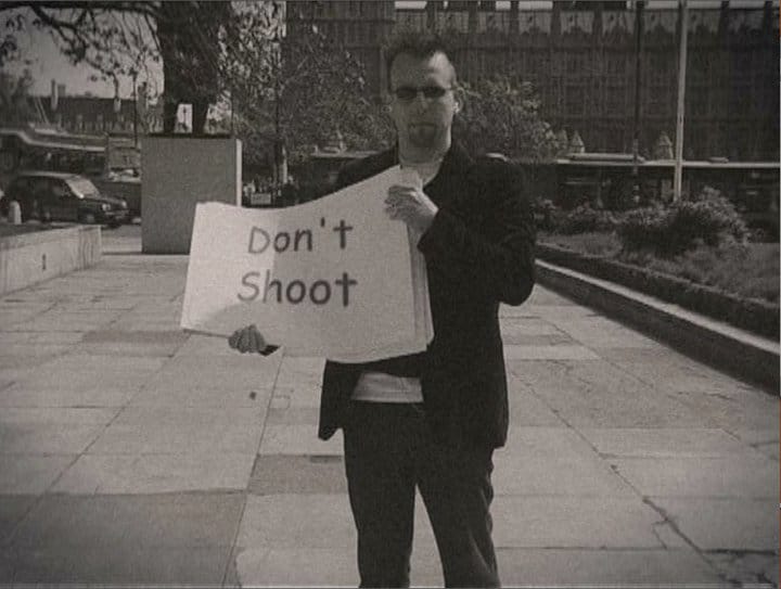 A screenshot from a documentary, the author in Parliament Square holding a blank protest placard with "Don't Shoot" digitally inserted onto it.