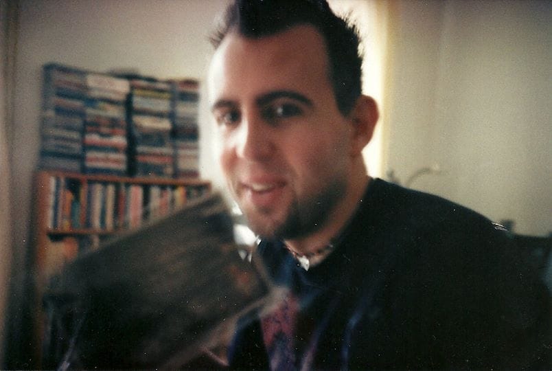 A blurry photo of me, a white person with dark hair, caught smiling, with a straining bookcase of books, CDs, and DVDs behind..