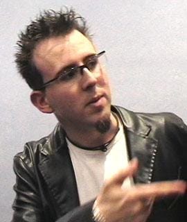 A white person with glasses, dark hair and goatee, wearing a white shirt and PVC jacket.