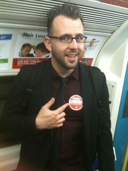 Me, a person with glasses, dark hair and stubble, on the London tube train pointing at a label that states "Hello, My Name is Jay Baker."