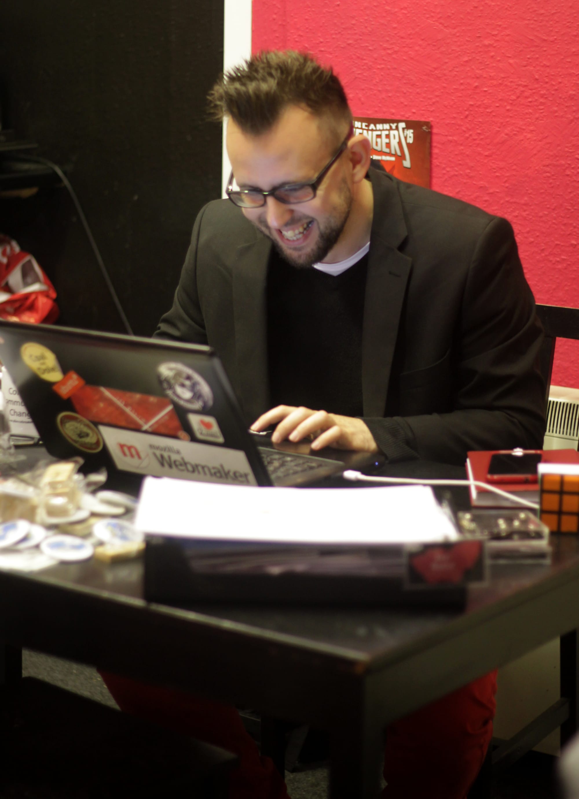 Me, a white person with glasses, dark hair and goatee, typing on a laptop while smiling shyly in the awareness of being photographed.