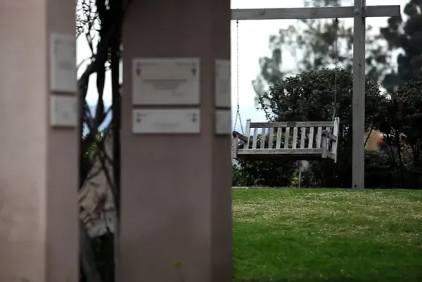 A wooden swinging bench from a distance across a grassy field. There are out of focus brown pillars in the foreground with dedication plaques affixed to the surface.