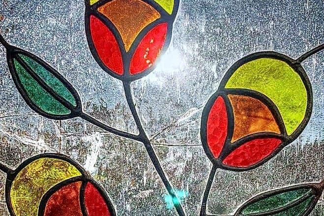 Stained glass of orange/yellow and red flowers with green leaves. Sun is shining though and there are cracks in the glass.
