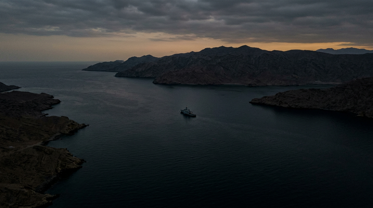 Empty Strait of Hormuz at dusk — a single naval vessel at the narrowest point, no commercial shipping.
