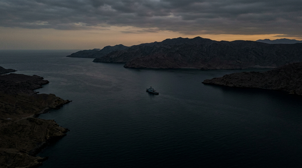 Empty Strait of Hormuz at dusk — a single naval vessel at the narrowest point, no commercial shipping.