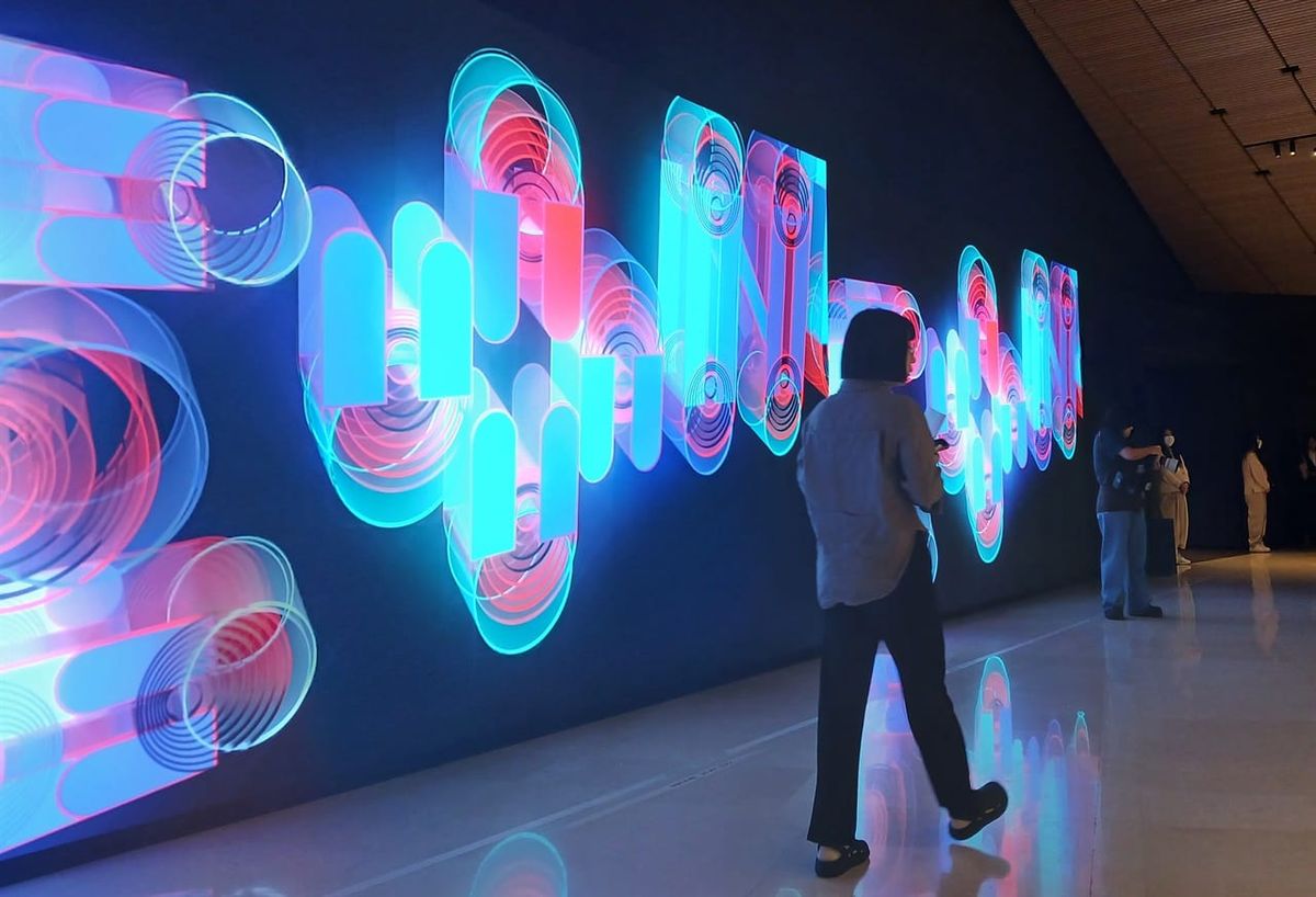 Eine Besucherin geht an einer langen LED-Wand in einem dunklen Museumsfoyer vorbei, auf der leuchtende, verschlungene Formen in Türkis und Rot zu sehen sind.