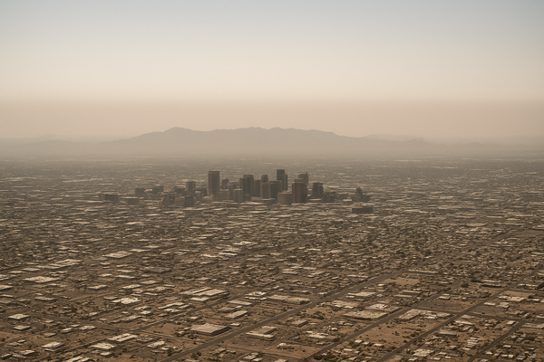 Wide aerial view of a hazy desert city skyline at midday, brown smog layer visible against a pale blue sky, no people