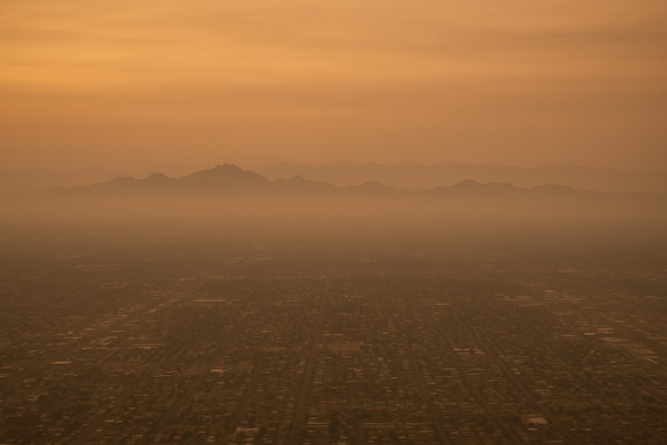 Wide aerial view of a hazy Phoenix valley at dusk, orange-tinted sky, mountain silhouettes in the distance, no people