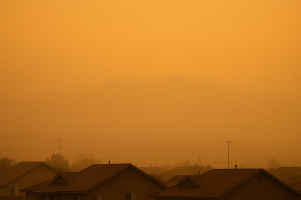 of a hazy desert sky at dusk over a suburban roofline, warm orange light diffused through thick air, no people
