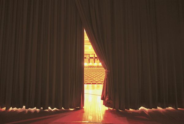 Empty concert hall stage with warm amber stage lighting, orchestra pit visible below, red velvet curtain partially drawn, no