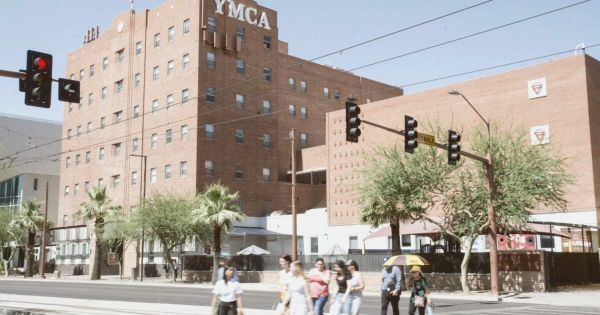 Exterior of an aging red-brick multi-story building on a quiet urban street corner at midday, desert sky above, no people