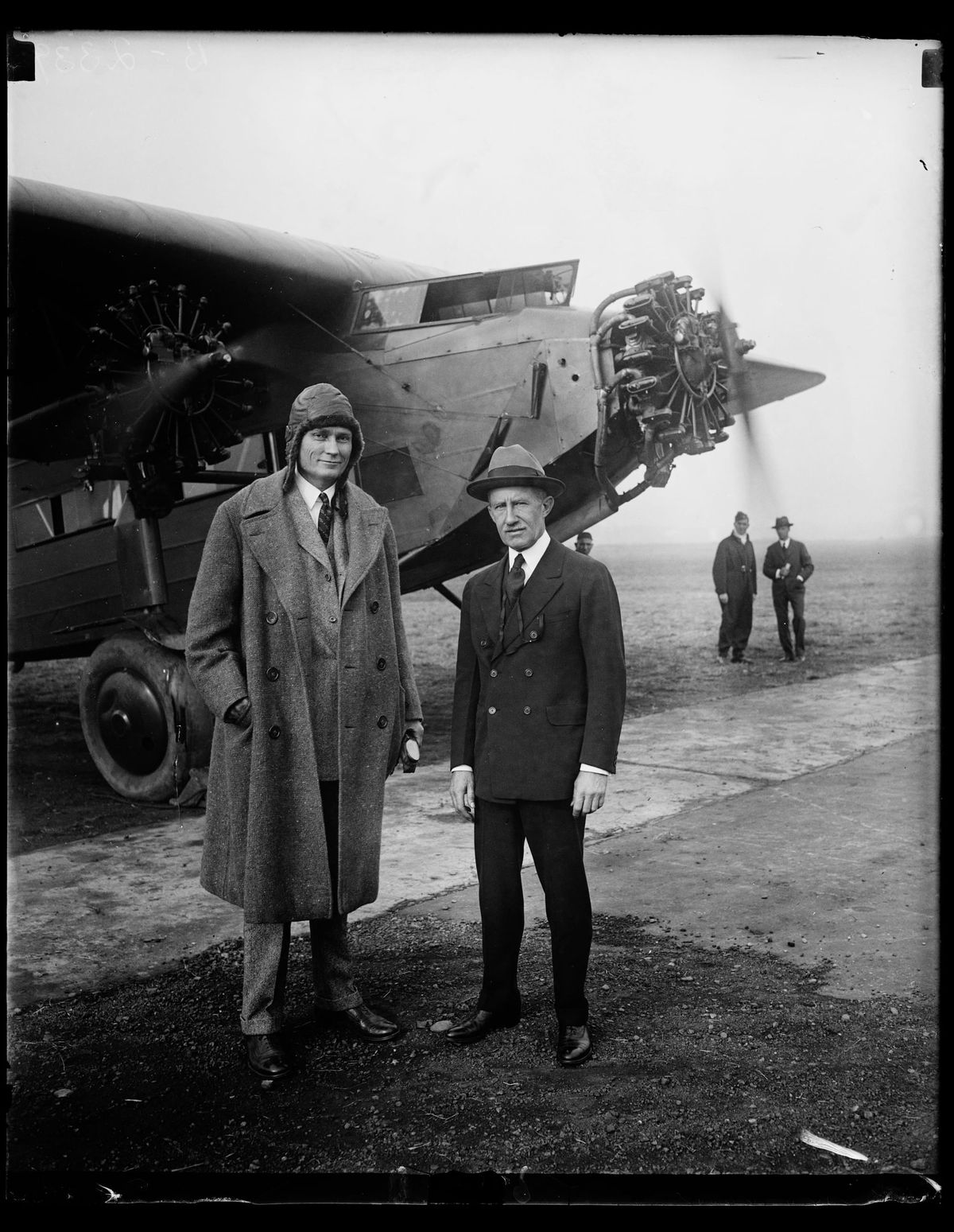 Two men stand in front of an airplane in a black and white photo from 1927. 
