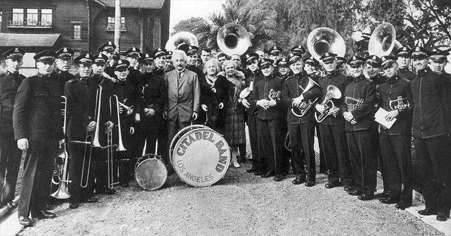 Black and white photo of Albert Einstein standing with a marching band dressed in black. 