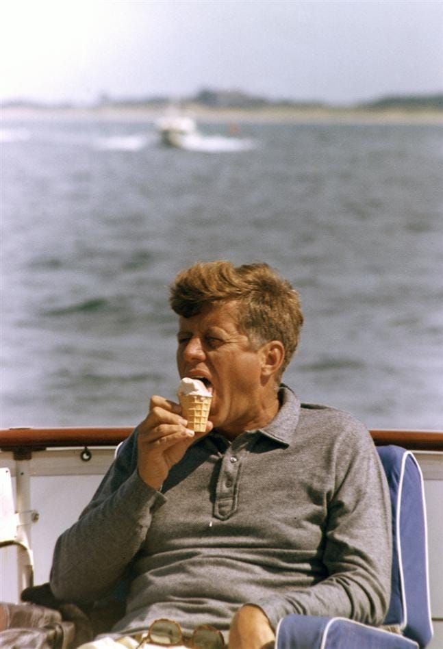 President Kenndy in a grey long-sleeve shirt sits in a boat, eating an ice cram cone. 