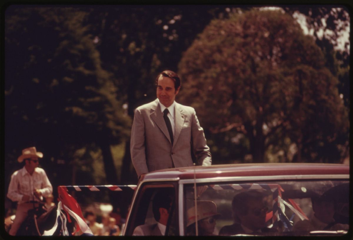Senator Bob Dole stands in the bed of a pickup truck wearing a suit; he is in a parade. 