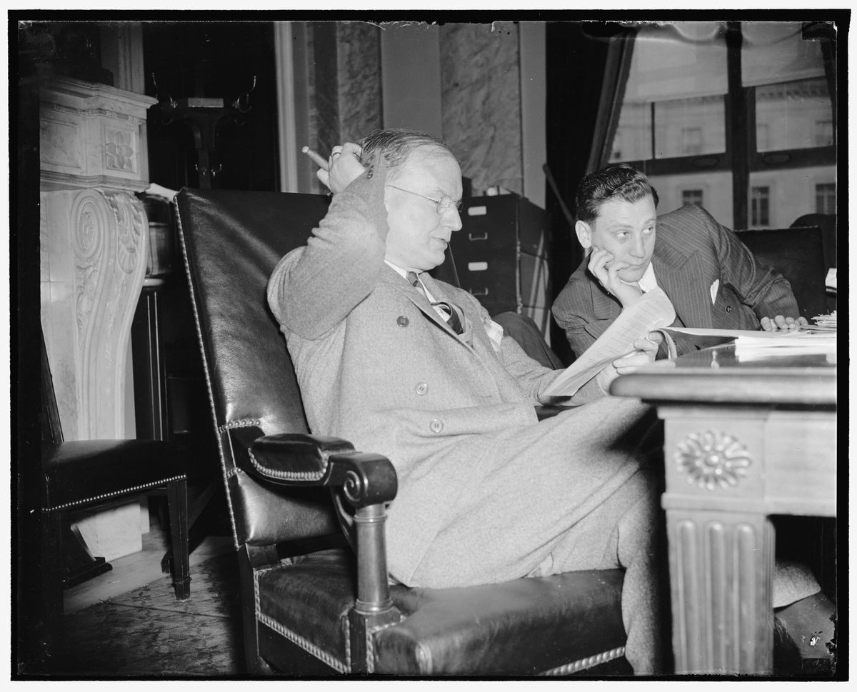 A black and white photo of two men, one with a cigar and glasses, sitting at a desk during a Senate Committee hearing.