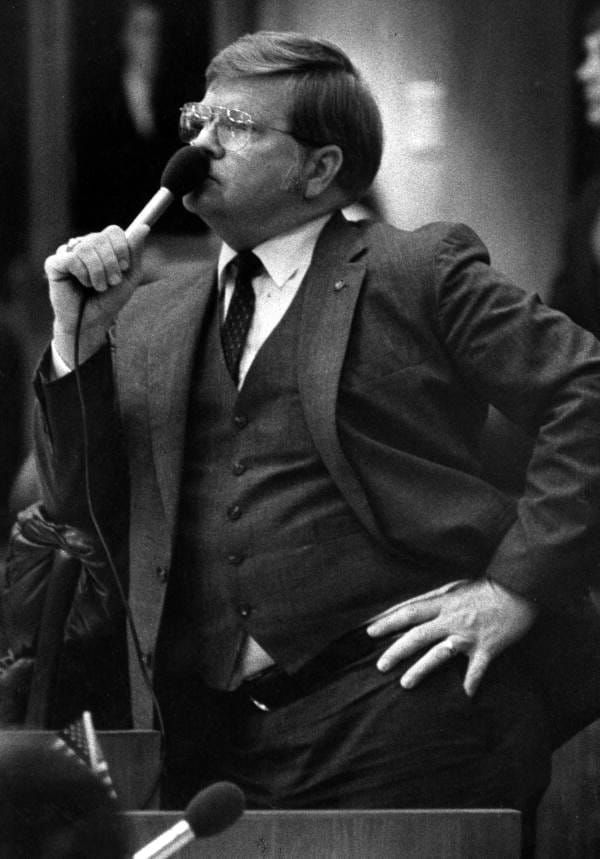 Black and white photo of a man wearing a suit, glasses, with his left hand on his hip, and his right hand holding a microphone very close to his face. 