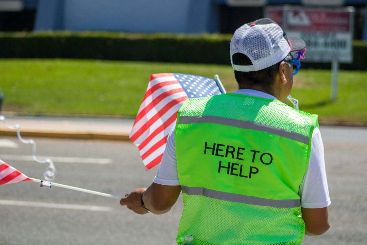 Man photographed from behind wearing a green vest that says, HERE TO HELP.