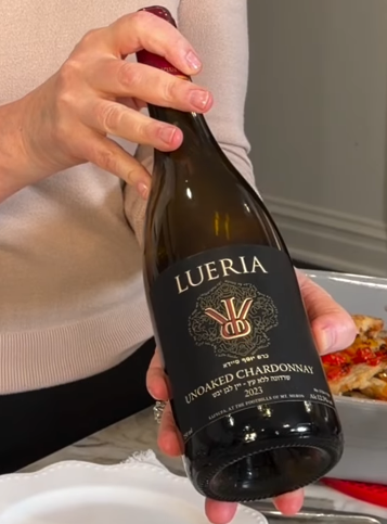 Anne holding a bottle of Lueria Unoaked Chardonnay 2023 with its black and copper front label, the pepper chicken in a baking dish behind it