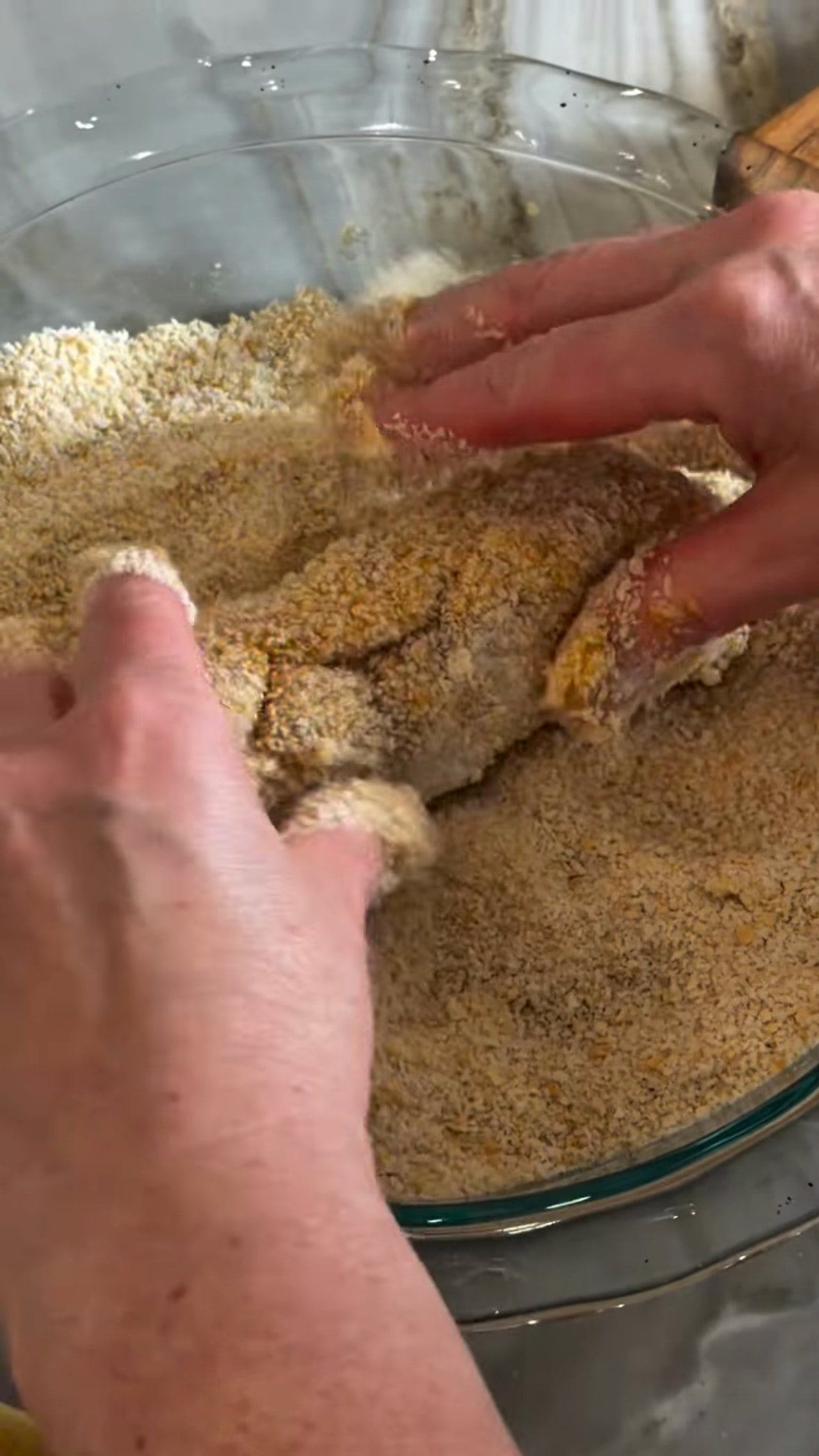 Hands pressing breadcrumbs onto a rolled chicken breast in a glass bowl