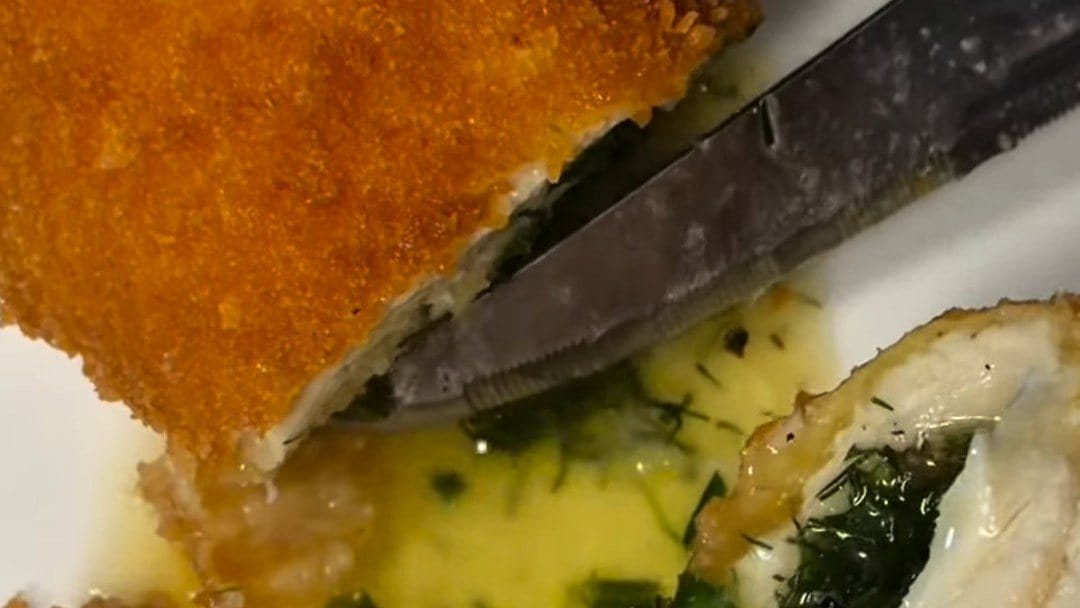 A knife slicing through golden, crumb-crusted chicken Kyiv as herb-flecked garlic butter spills onto the plate
