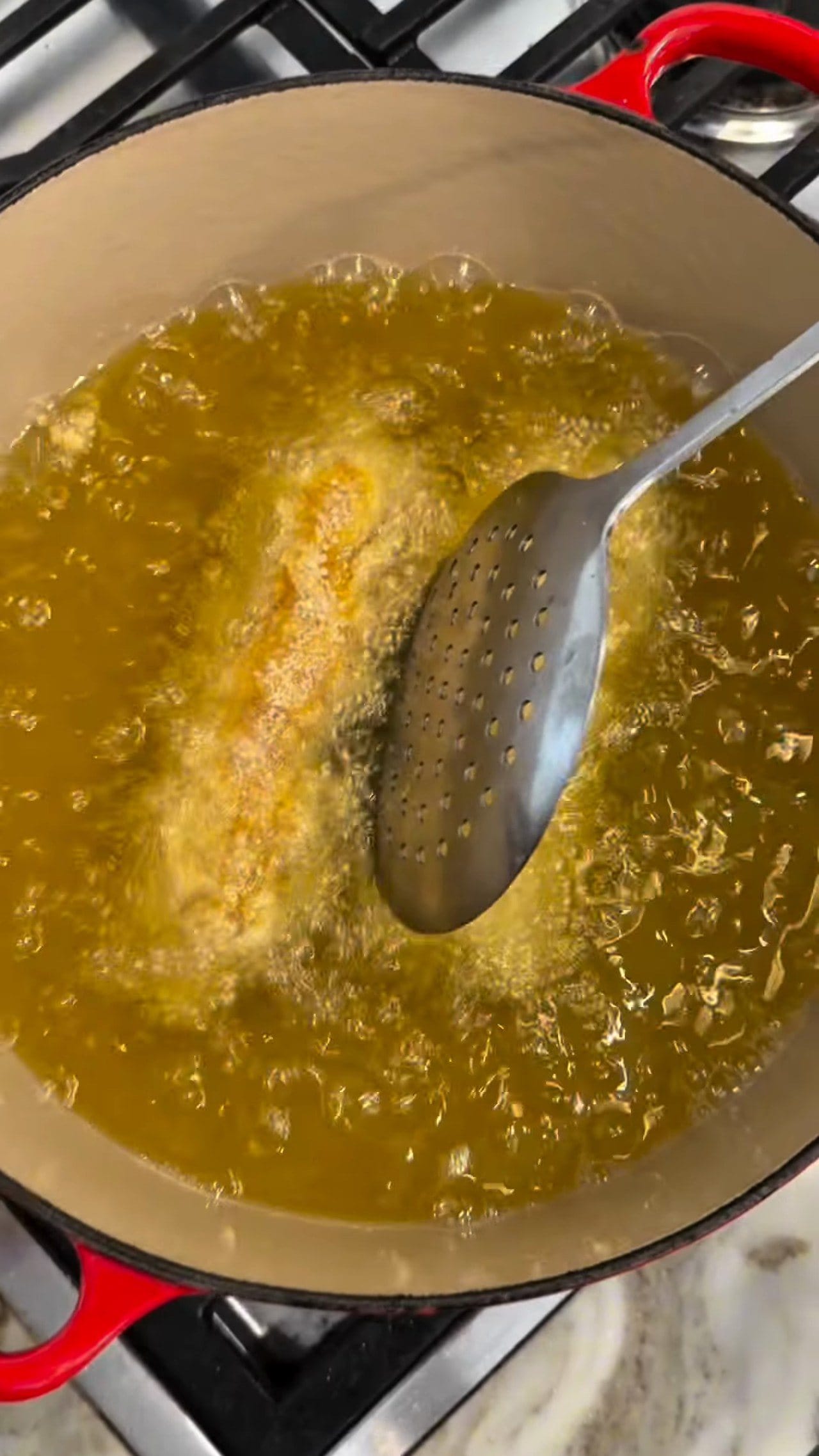 A breaded chicken Kyiv frying golden in bubbling oil in a red enameled pot, turned with a slotted spoon