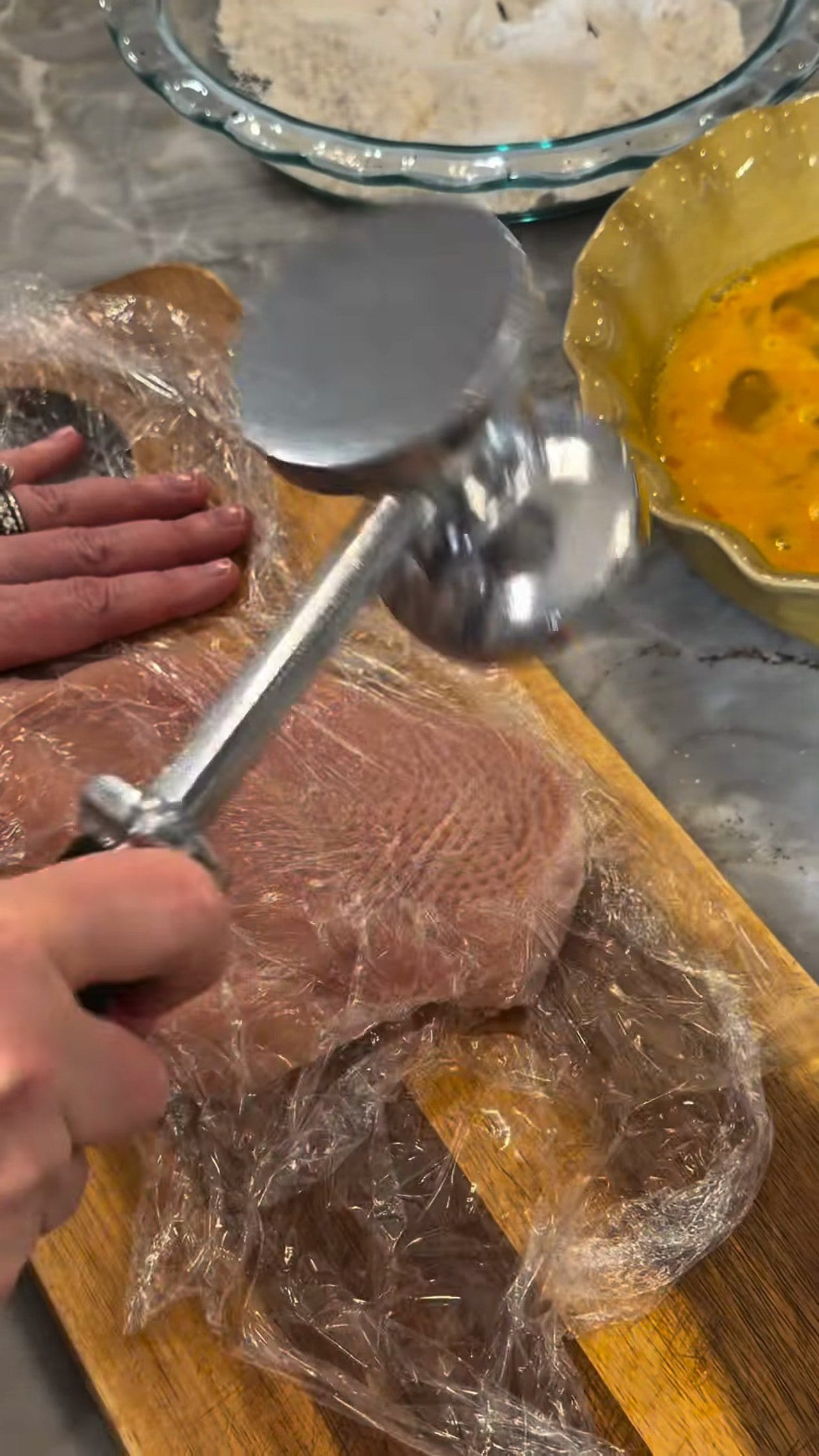 Anne pounding a chicken breast thin under plastic wrap with a meat mallet, bowls of flour and beaten egg waiting behind it