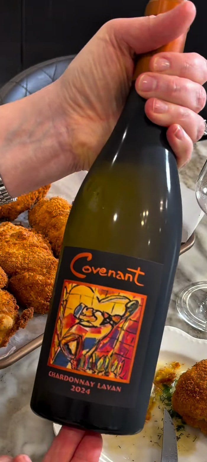 Anne's hand holding Covenant Chardonnay Lavan 2024, its painted label framed in orange, beside a tray of golden chicken Kyiv