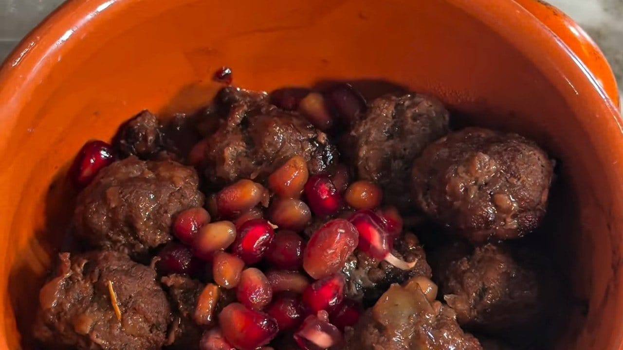 Close-up of browned meatballs in glossy pomegranate sauce, topped with fresh pomegranate seeds in an orange bowl