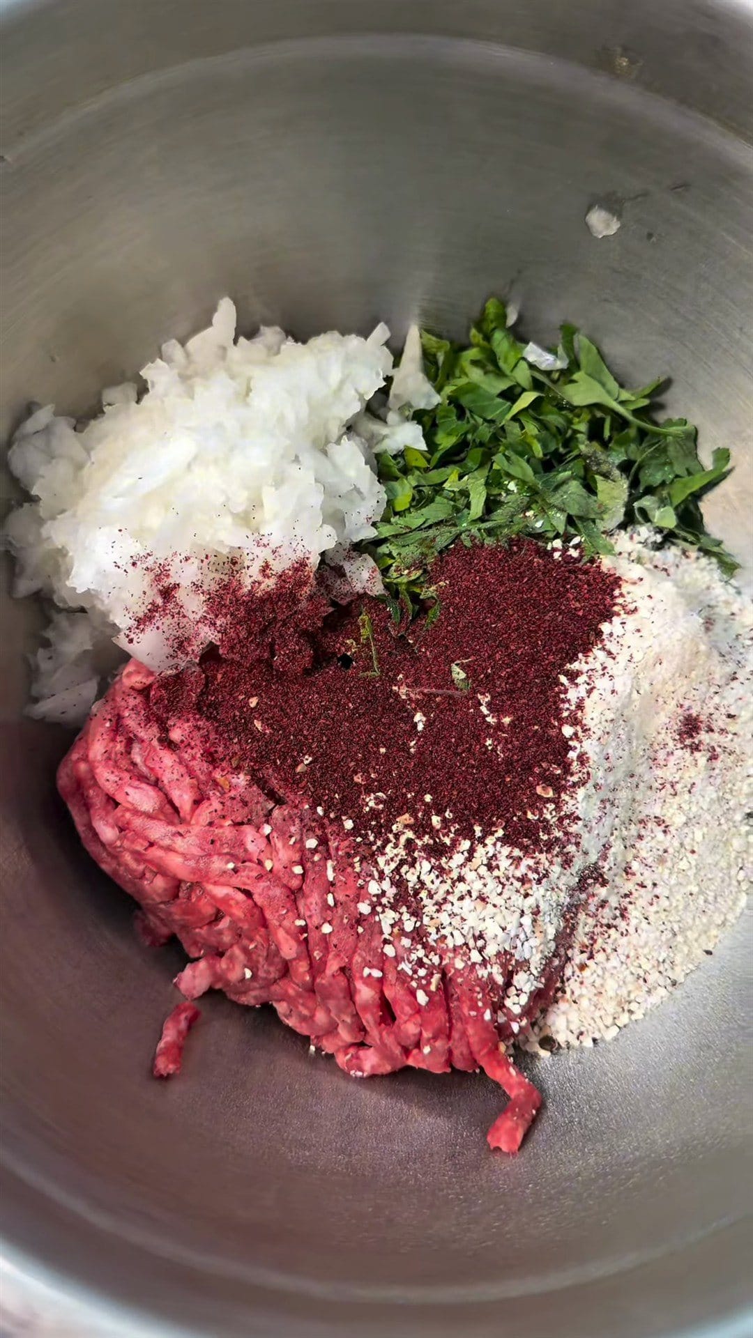 Ground beef, grated onion, chopped green herbs, breadcrumbs and a dark red spice in a steel mixing bowl
