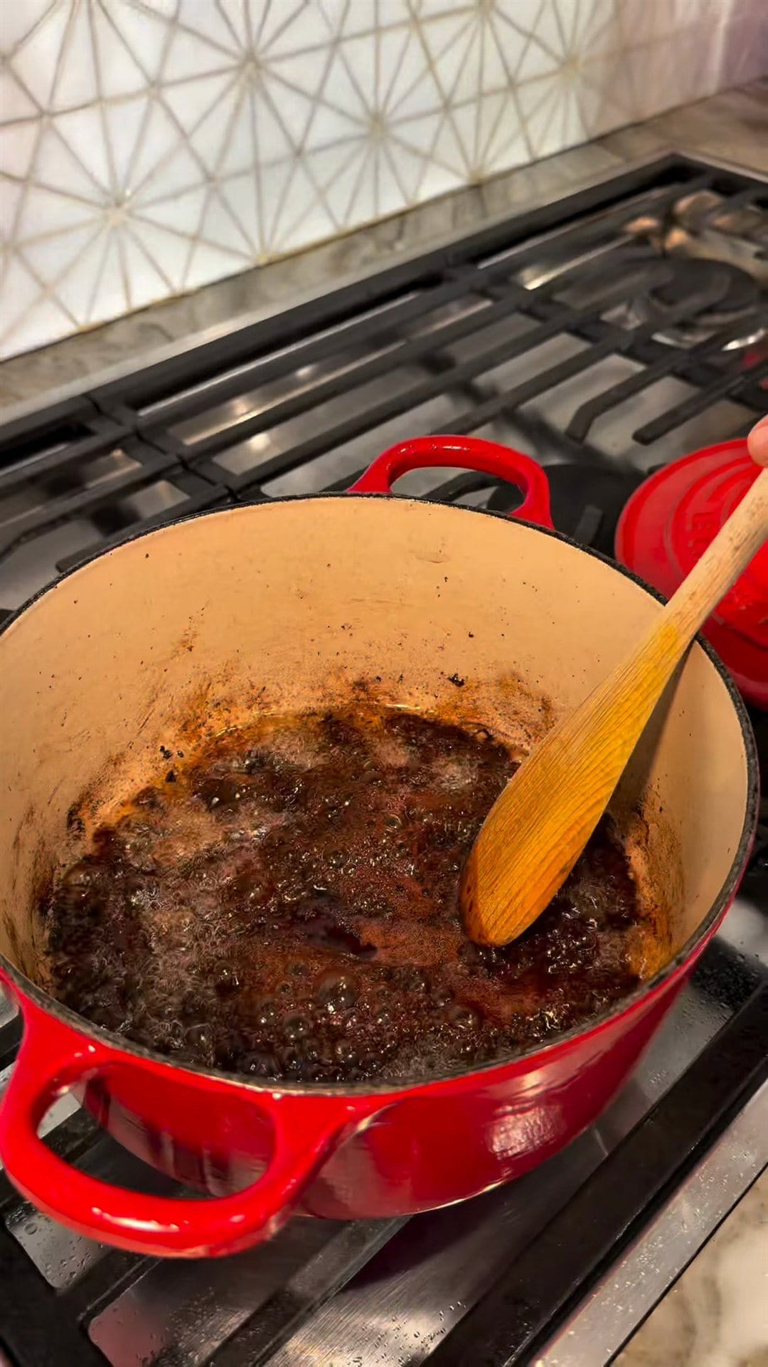 A wooden spoon stirring dark, bubbling pomegranate sauce in the red Dutch oven