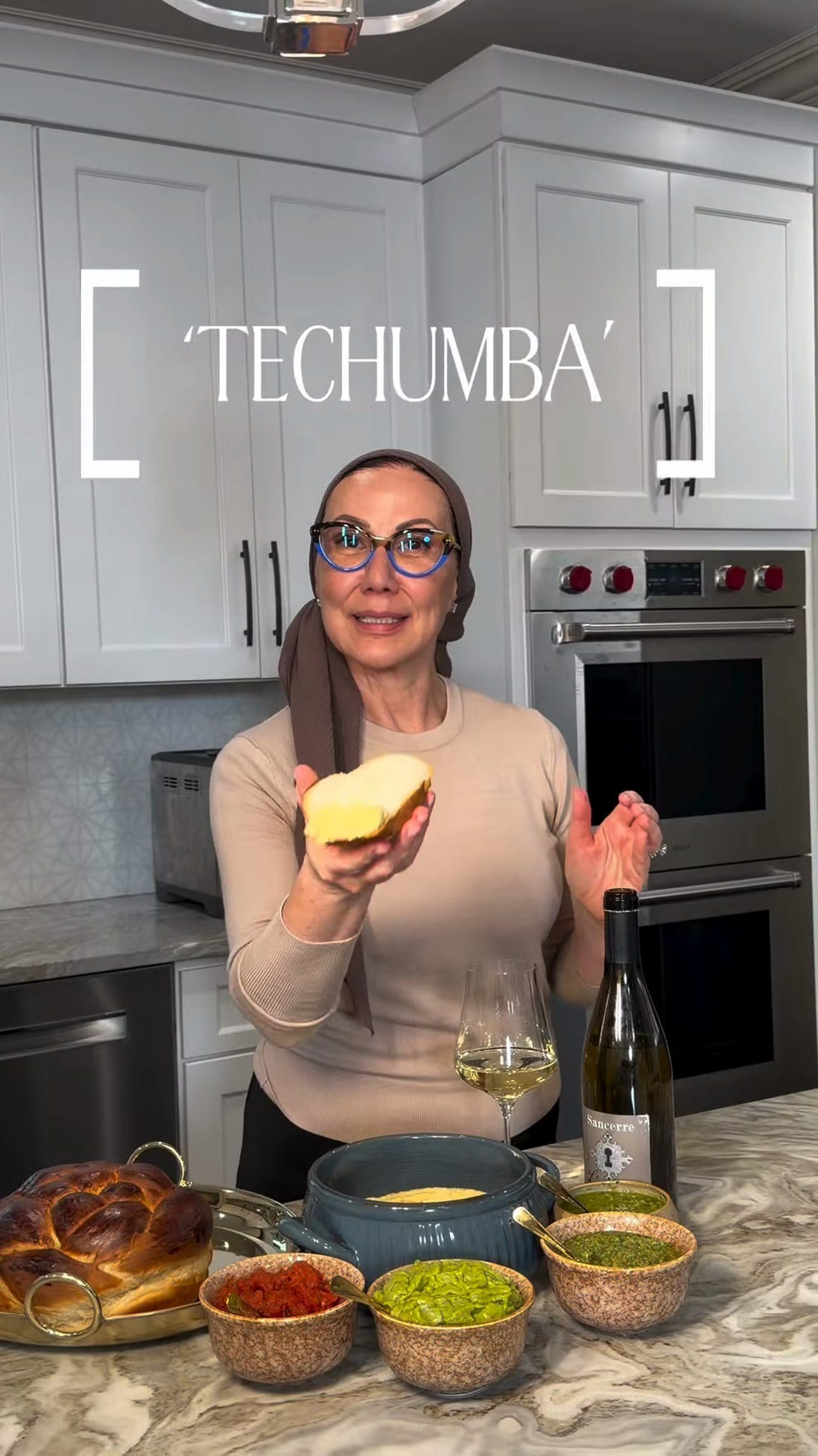 Anne in her kitchen holding challah above five bowls of dips and a bottle of Sancerre, with the title Techumba