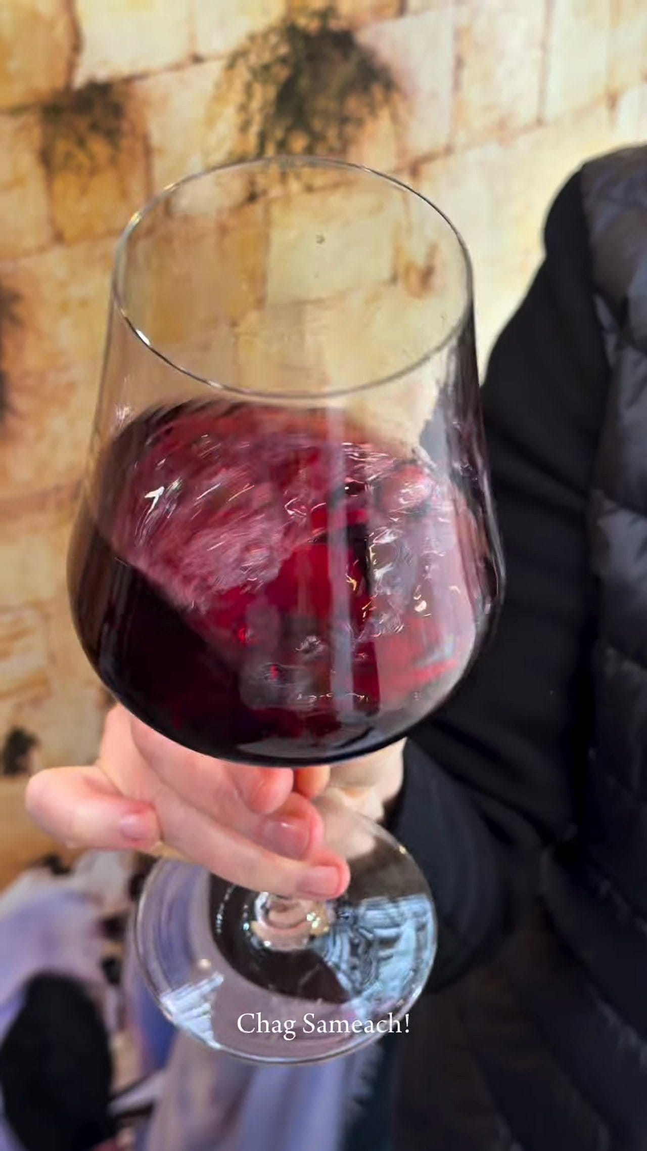 A glass of red wine held up in the sukkah against the Western Wall mural
