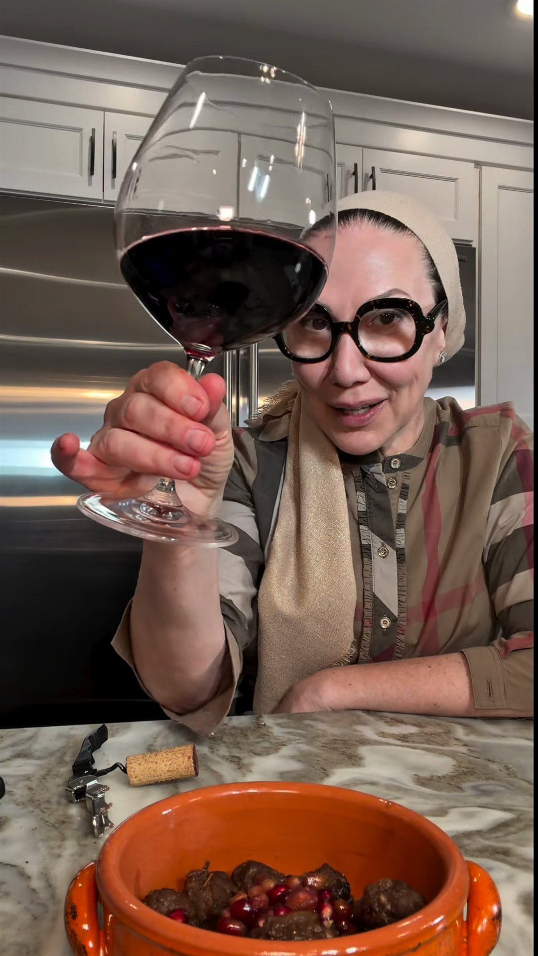 Anne at her kitchen counter raising a glass of red wine over a bowl of pomegranate meatballs, a cork and corkscrew beside it
