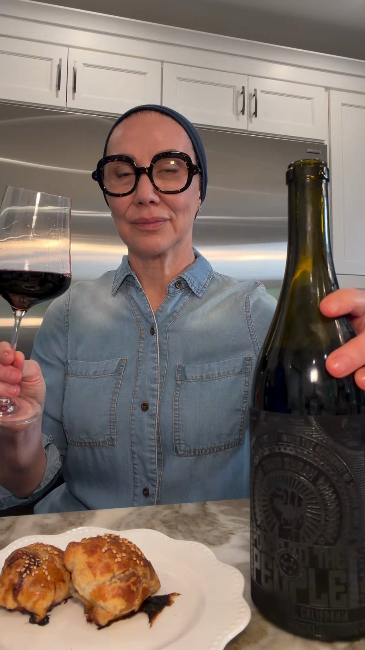 Anne at her kitchen counter with a glass of Shirah Syrah, the bottle and a plate of golden samsa