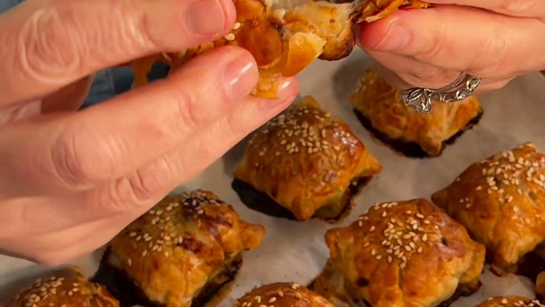 Hands pulling apart a flaky samsa over a tray of golden, sesame-topped samsa