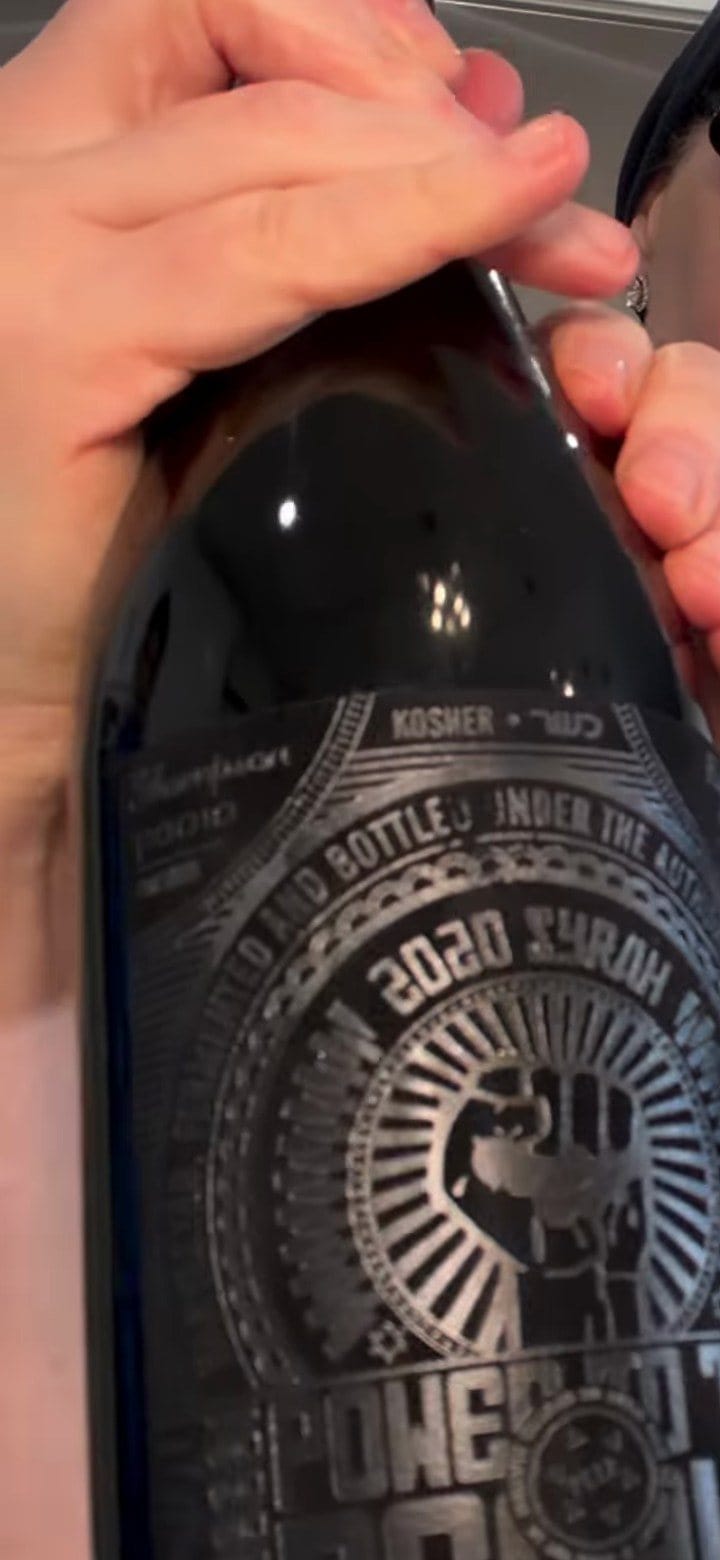 Anne holding the Shirah Power to the People 2020 Syrah, its dark label marked Kosher with a raised-fist emblem