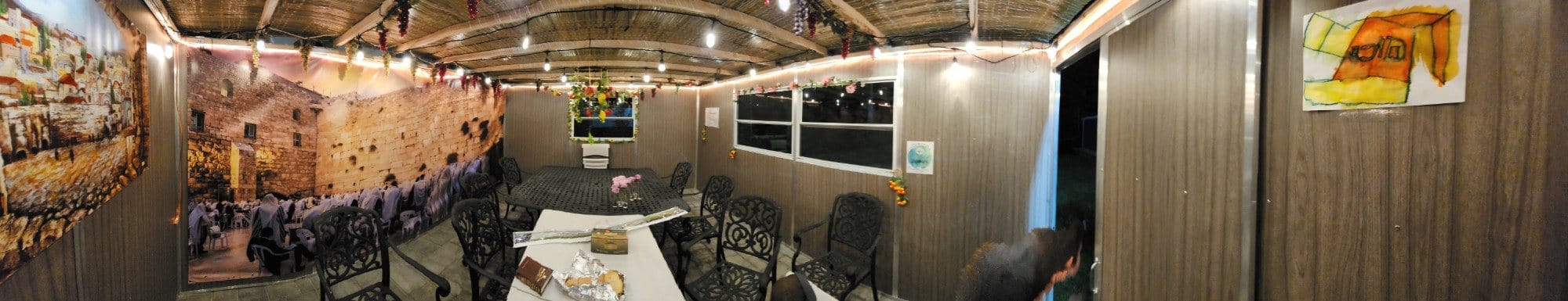 Panoramic view inside a decorated sukkah at night: a long table with wrought-iron chairs, string lights and grape clusters hanging from the schach, a Western Wall mural and a child's drawing on the walls