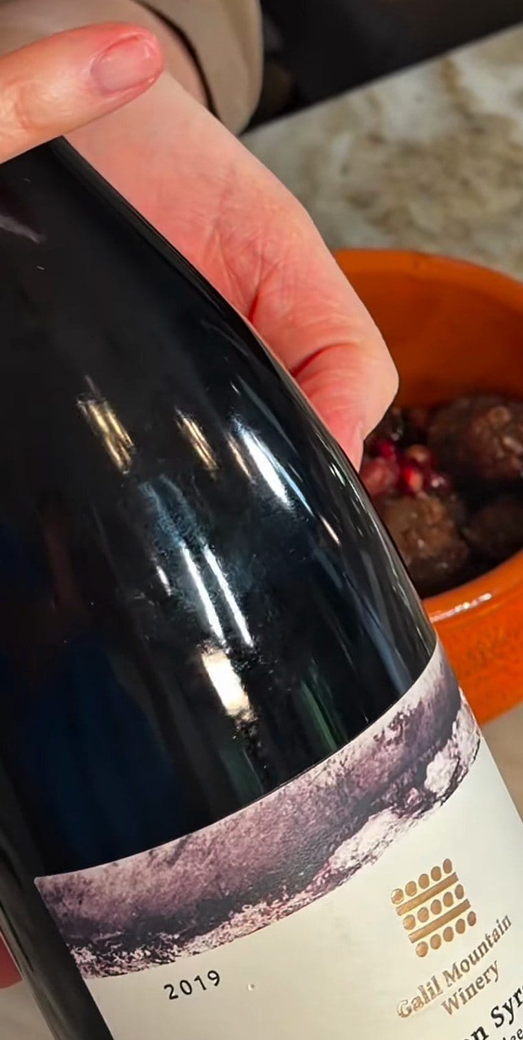 Anne's hand holding a 2019 Galil Mountain Winery Yiron Syrah, with the bowl of pomegranate meatballs behind it