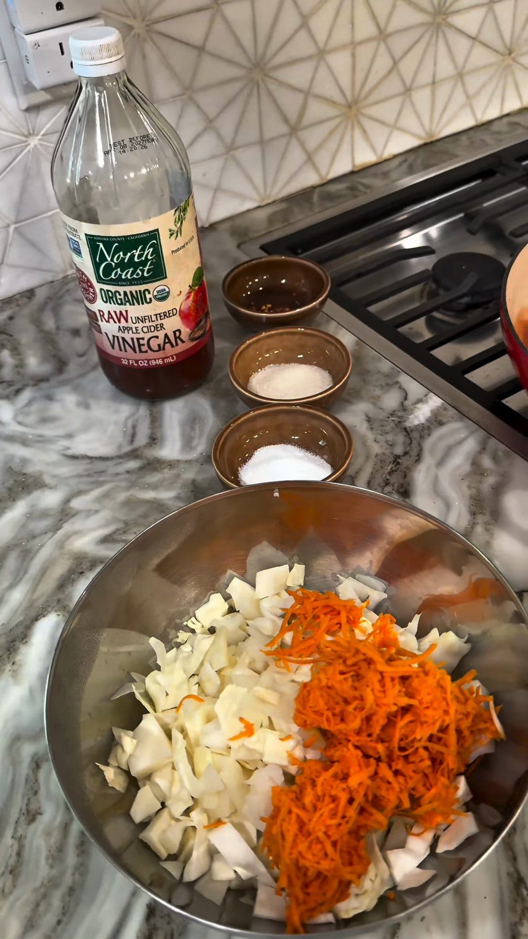 Steel bowl of chopped white vegetables and grated carrot, small bowls of seasonings, and a bottle of apple cider vinegar by the stove